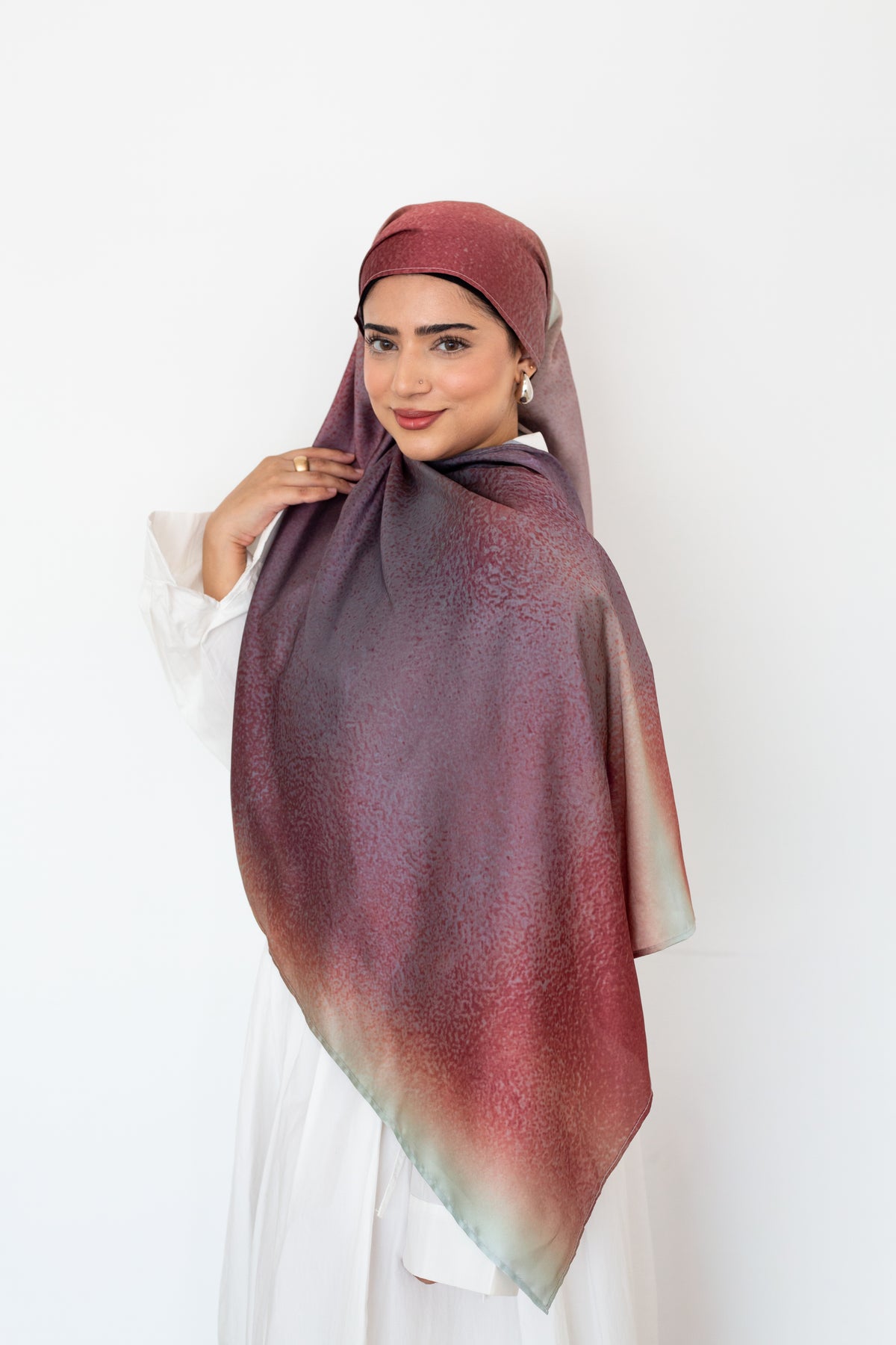 A woman wearing a hijab with a gradient color transition from deep burgundy to muted green, paired with a black undercap, a white button down and a matching white skirt against a plain white background.