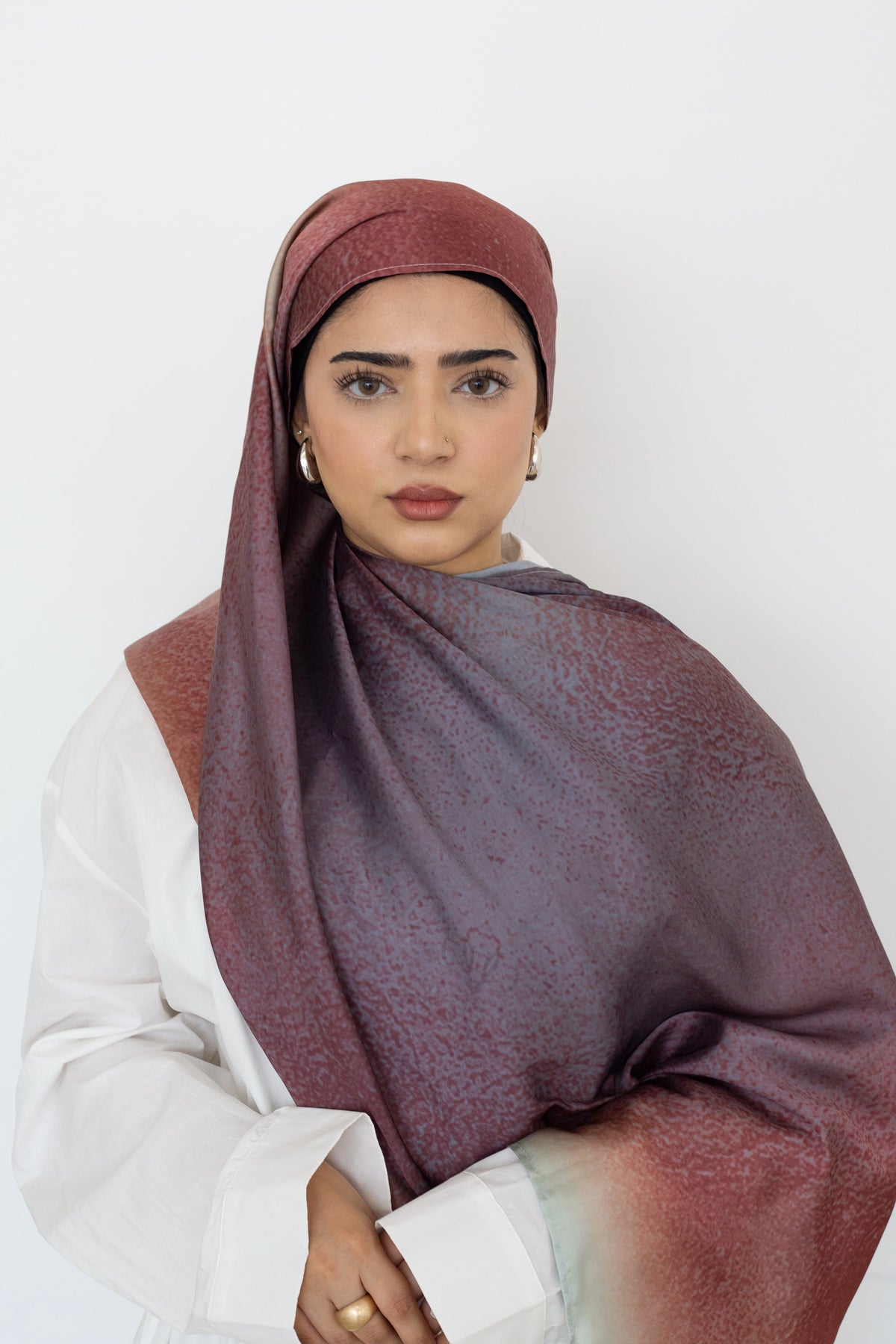 A woman wearing a white button down and a large draped hijab or shawl in a dusty mauve-purple color with a subtle textured pattern and a reddish-brown gradient at the edges, set against a plain white background.