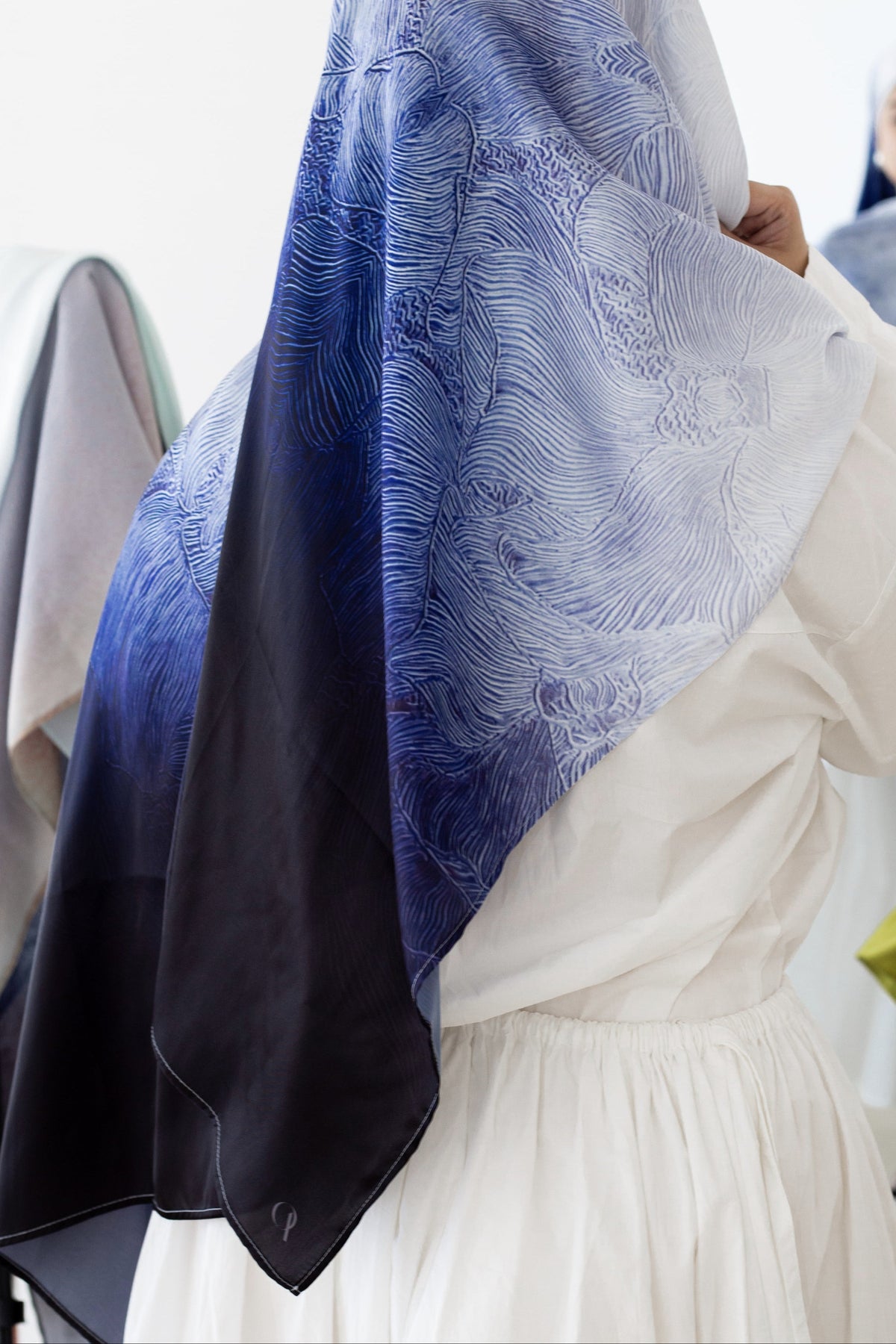 A person wearing a white dress and a large patterned scarf with a blue-to-black ombré gradient, shown from behind in a bright indoor setting.