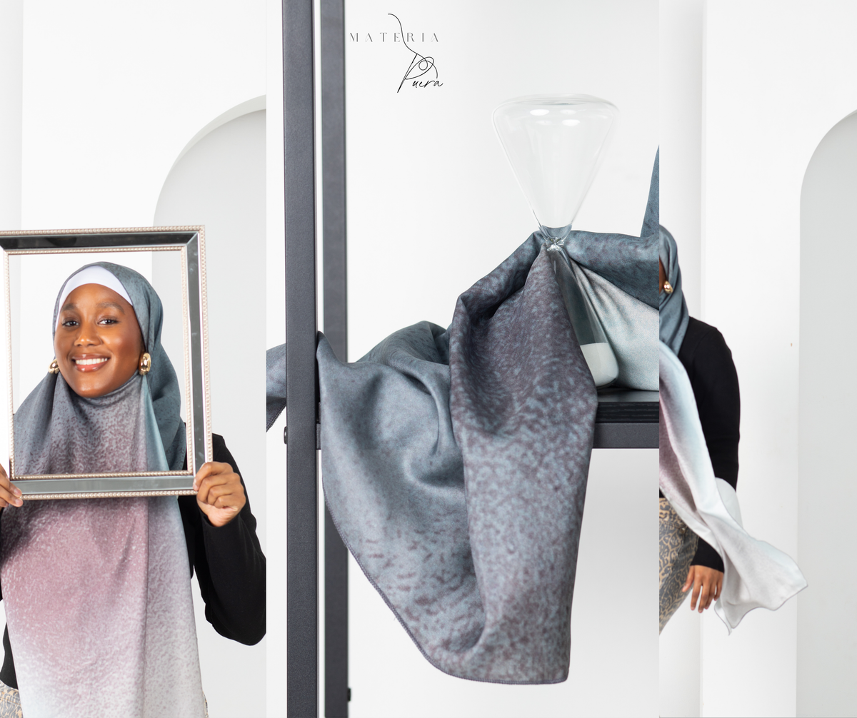 A woman wearing a patterned hijab with a gradient of sky blue, mauve, and petal pink, posing with a picture frame on the left, while draped fabric of the same hijab hangs from a black stand on the right. The background is a clean white studio space with arches.